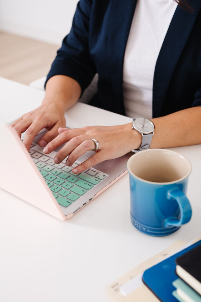 Hands typing on laptop with blue mug beside.